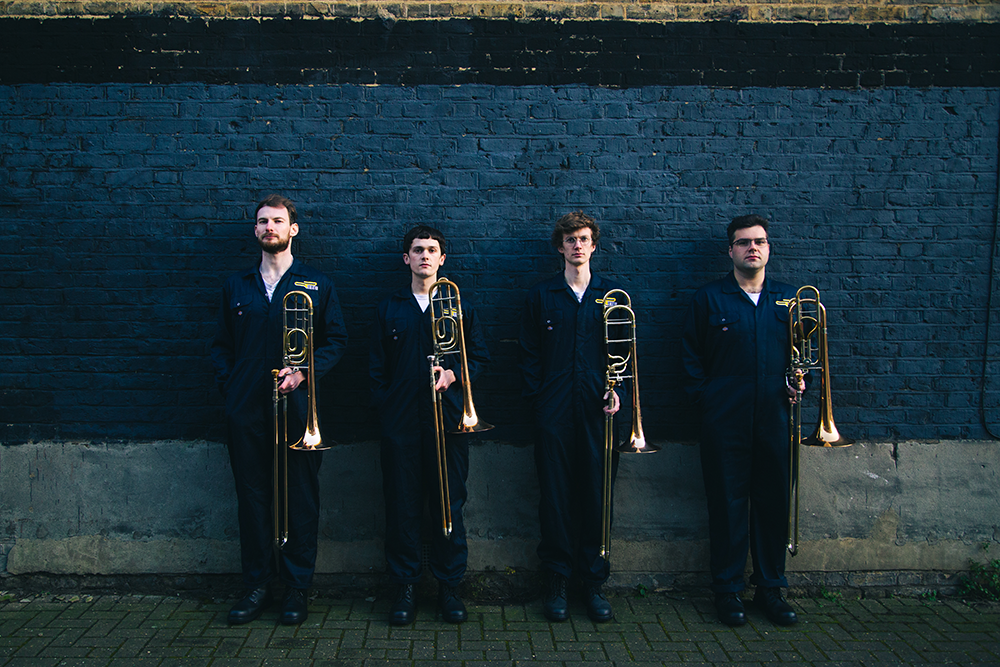 The four members of Slide Action stand dressed in blue boilersuits, upright against a blue wall, their trombones by their sides